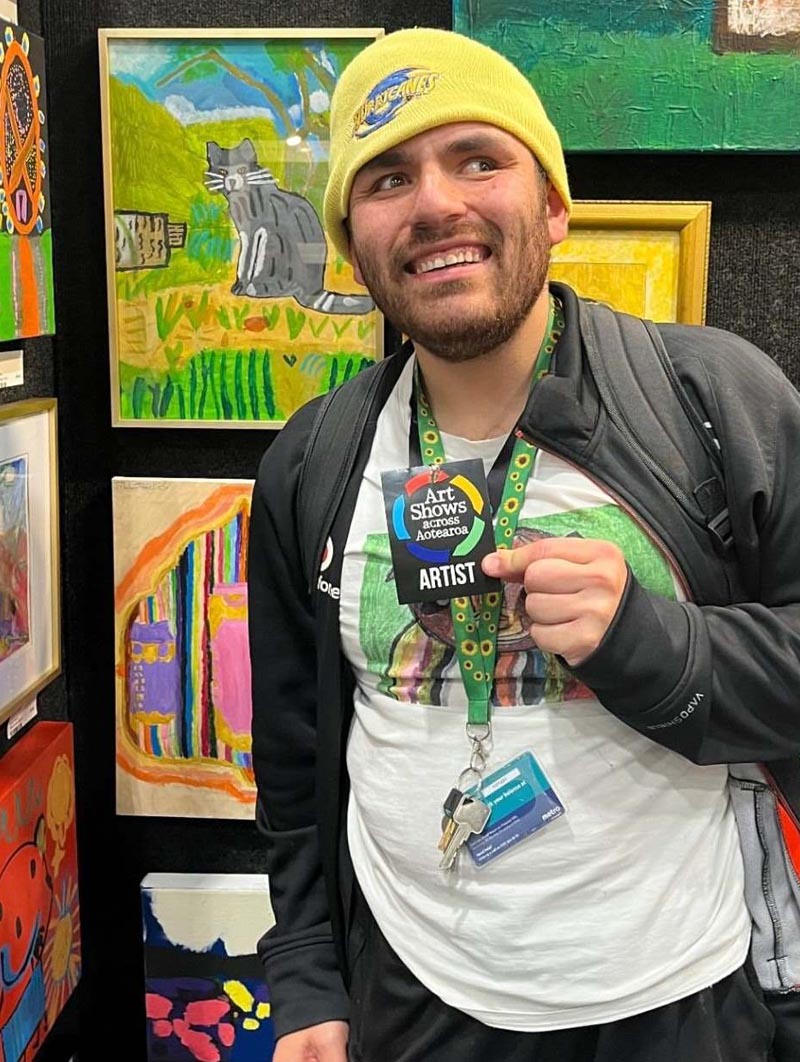 A smiling Jagar Sowman is holding a lanyard from the Art Shows with the label Artist on it. He is surrounded by paintings hanging on the wall.